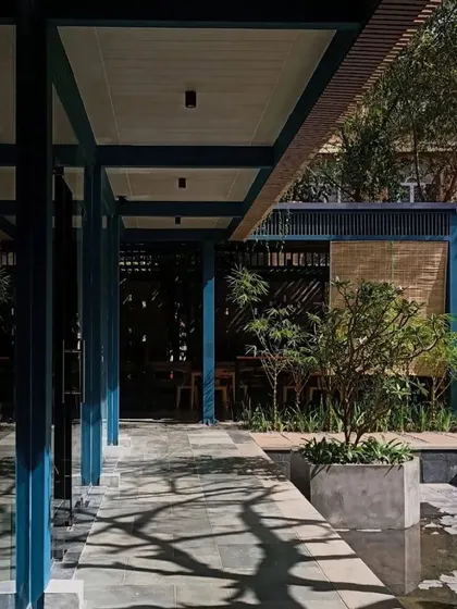 The main walkway at IRIS Cafe, where shadows from the trees dapple the path. The use of blue steel columns adds a contemporary touch to the otherwise natural and earthy palette.