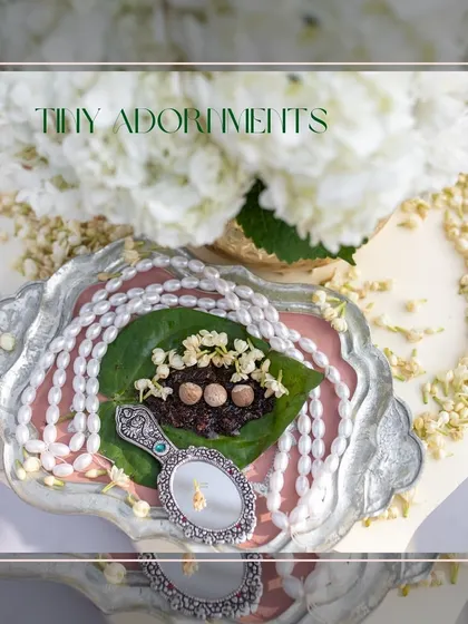 Tiny adornments for a welcome ceremony, featuring a betel leaf with supari, a pearl necklace, and a vintage hand mirror on a silver tray.