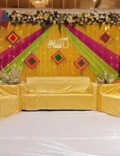 A stage setup for a Haldi function, featuring a backdrop of yellow, green, and pink drapes, floral arrangements, and a modern neon "Haldi" sign.