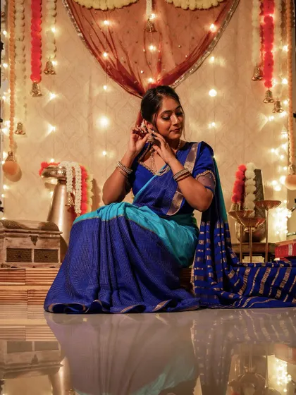 My Deepavali look. I'm sitting amidst a festive setup of diyas and flowers, wearing a beautiful dual-tone blue Mysore silk saree. The traditional decor perfectly complements the festive vibe of the outfit.