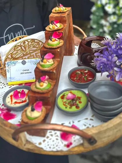 Chana Jor Garam, elevated. We serve this classic street snack on crisp mathris with fresh toppings and a duo of chutneys, all presented on an elegant wooden tray adorned with flower petals.