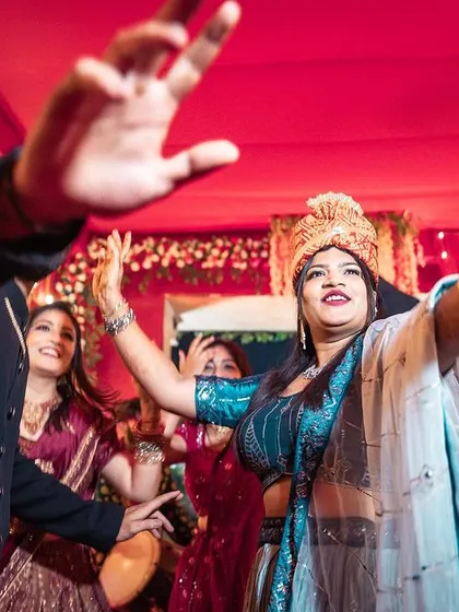Capturing the bride's grand entrance with her hands up, fully embracing the music and celebration. The dynamic angle and vibrant red background amplify the festive atmosphere of this candid wedding moment.