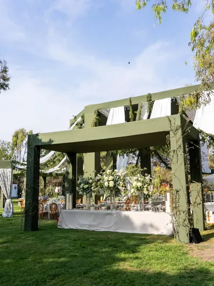 A wide shot of a food buffet station set under a large green pergola. The table is decorated with grand arrangements of white flowers, ensuring the dining area is as beautiful as the rest of the venue.