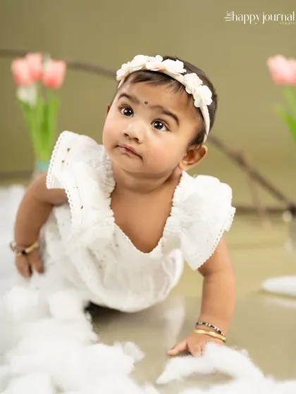A baby girl in a white romper crawls through a set of soft clouds, looking up with curiosity. A creative and adorable setup.