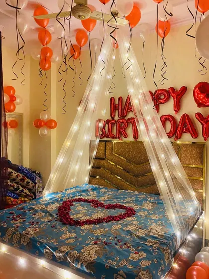 A classic romantic birthday setup with a fairy light canopy over the bed. The room is decorated with red and white balloons, and a heart of rose petals on the bed adds a special touch.