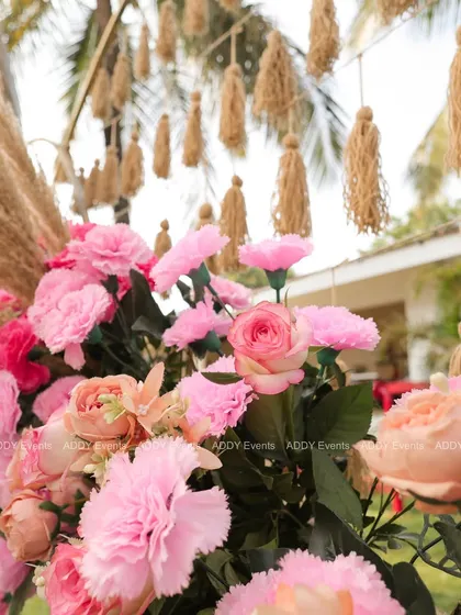A close-up shot of the floral decor on a Mehndi arch, showing the beautiful combination of pink carnations, peach roses, and fluffy pampas grass. In the background, hanging jute tassels add to the rustic, handcrafted feel.
