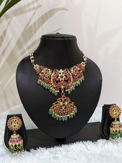 An intricate traditional Jadau choker with a floral pendant, paired with matching jhumkas. The ruby and emerald stones with strawberry bead drops make this a colorful and festive choice.