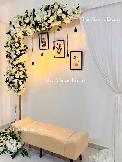 A minimalist and modern engagement setup. This design features an L-shaped floral stand with white flowers, paired with hanging Edison bulbs, framed pictures, and a simple bench for a chic and personal touch.