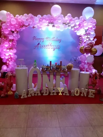 A classic pink-themed first birthday for Aradhya. The setup includes a beautiful balloon arch, a simple backdrop, and large "ONE" letters, creating a grand focal point in the party hall.