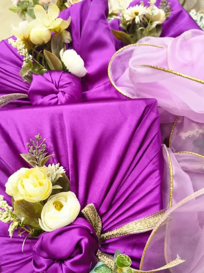 Another angle of the Furoshiki packaging for an engagement ceremony. The rich purple fabric is gathered and tied with a gold ribbon, then decorated with small yellow and white flowers.