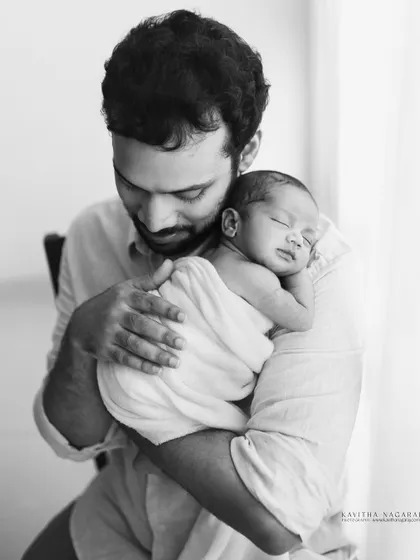A father holds his sleeping newborn, swaddled in white. This black and white portrait, taken near a window, is full of soft light and quiet emotion.