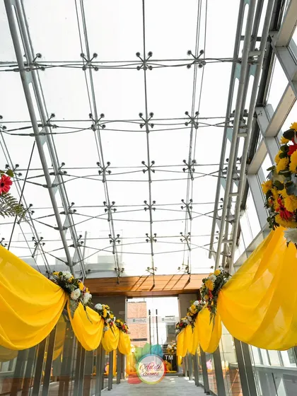 A sunlit glass-covered walkway decorated with yellow drapes and floral bouquets for the Haldi ceremony.