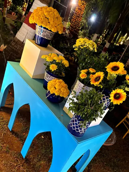 A corner display table in bright blue, adorned with an assortment of blue and white pottery filled with marigolds and sunflowers.