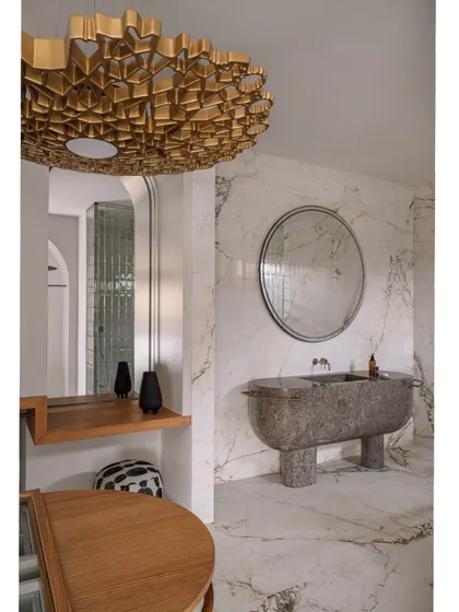 A full view of the monolithic granite basin, which stands like a sculpture against the white marble walls. The round mirror and gold light fixture complete the look.