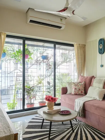 A homey feel is achieved by layering textures and patterns. In this living room, a plush pink sofa, a geometric rug, and a gallery wall of plants by the window come together to create a lived-in, eclectic vibe.