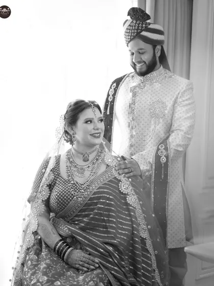 A classic black and white wedding portrait. The groom stands behind the bride, his hand on her shoulder, as they share a gentle moment. The monochrome tones add a timeless, elegant feel.
