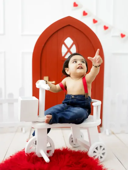 A playful moment on the tricycle, with the baby pointing up in excitement. The simple set design keeps the focus on his joyful expression.