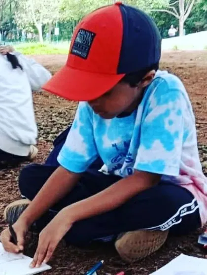 A child sits on the ground, fully absorbed in drawing in his nature journal during our "Tree Loving Tuesday" at the park.