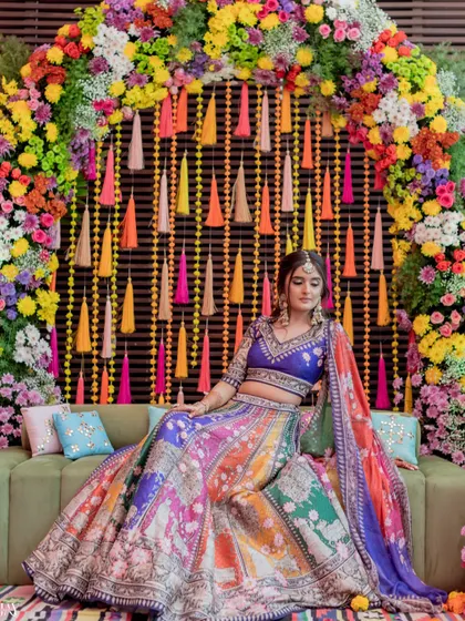 The bride looking regal in her multi-colored lehenga, seated against a vibrant floral arch.