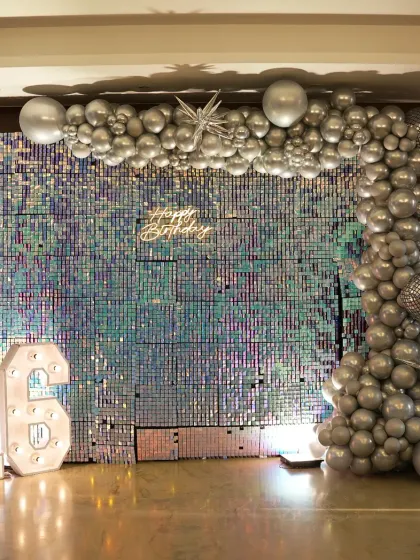 A dazzling setup for a 16th birthday party. The iridescent sequin wall creates a stunning, shimmering backdrop, complemented by a silver balloon garland and large marquee numbers.