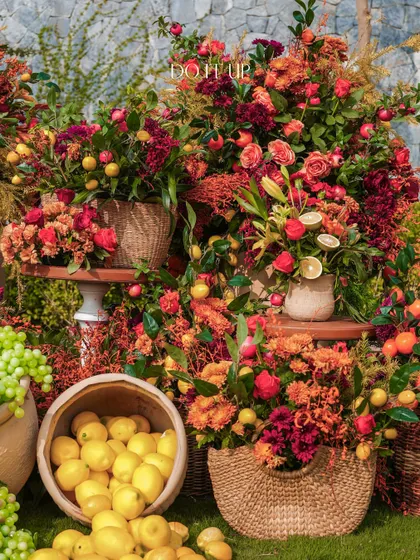 A beautiful, overflowing installation of flowers and fruits. The mix of red and orange blooms with fresh lemons and grapes creates a look of abundance and rustic charm, perfect for a daytime event.