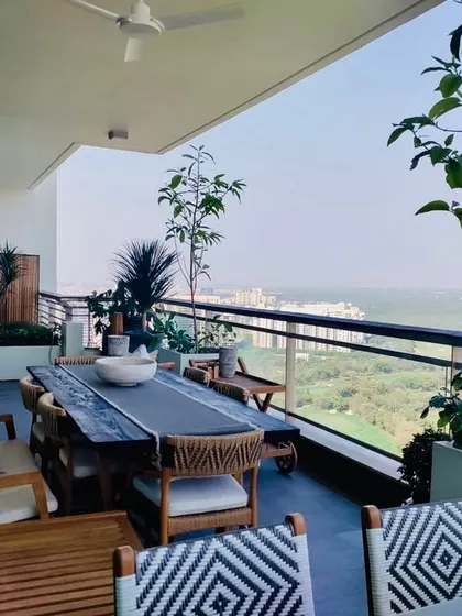 A full view of a spacious balcony designed for entertaining. It features a long dining table, woven chairs, and planters filled with trees and shrubs, all overlooking a stunning view.