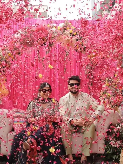 A couple showered in pink rose petals during their Mehendi, seated against a vibrant pink floral backdrop.