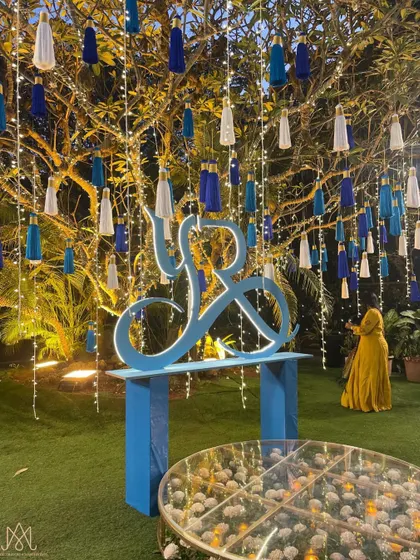 A personalized corner featuring the couple's monogram in a vibrant blue, set under a tree adorned with hanging tassels and fairy lights.