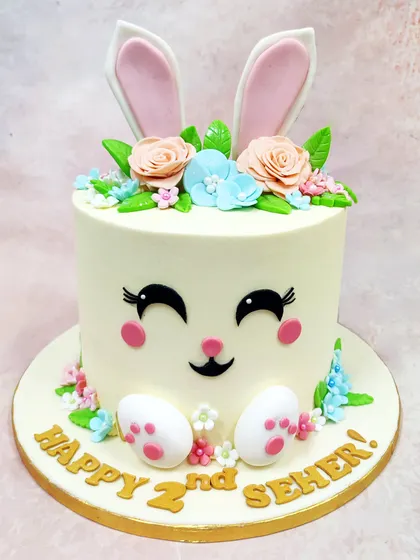 We craft pure pastel joy with this adorable bunny face cake. Perfect for a second birthday, this design features a sweet bunny face with large ears peeking out from a crown of delicate, handcrafted sugar flowers in soft peach and blue.