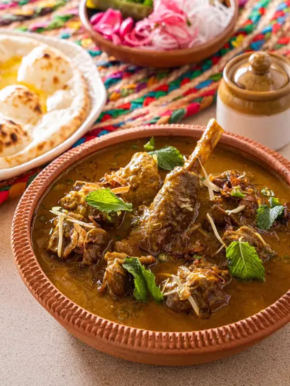 A rich lamb curry in a traditional terracotta bowl, served with naan. The styling emphasizes the rustic and authentic nature of the cuisine.