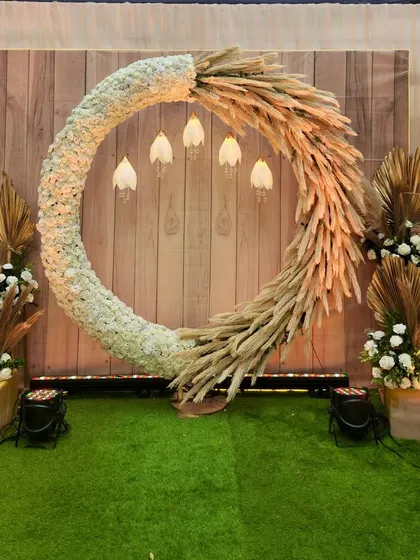 A beautiful bohemian photo booth with a large circular frame made of white flowers and pampas grass. The wooden backdrop and hanging lights add to the rustic, earthy charm.