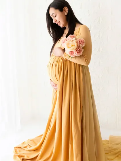 A full-length portrait of a mother-to-be in a long yellow gown, holding a bouquet of flowers. The soft, natural light from the window enhances the gentle mood of this boho session.
