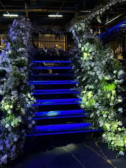A staircase at an engagement party, beautifully decorated with cascading white floral arrangements and illuminated with dramatic blue lighting.