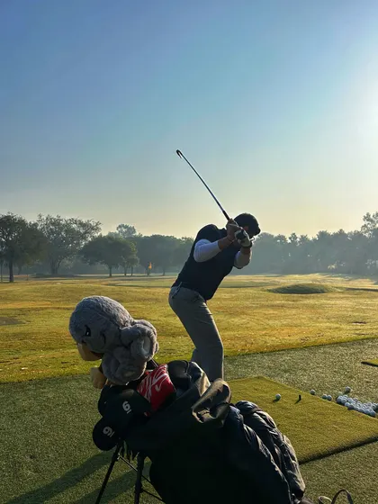 A view over a golf bag with a fun headcover, showing a golfer in mid-swing against the morning sun.