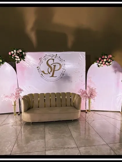 A chic and modern reception stage with a minimalist aesthetic, featuring three white arched panels, a custom gold monogram, and a stylish beige sofa, accented with simple floral bouquets on tall stands.