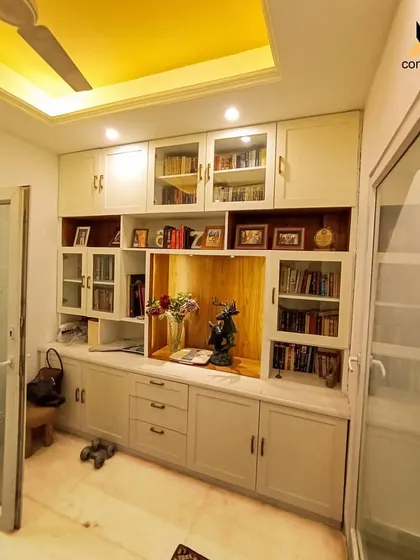 A full view of the custom-built library and storage unit. This piece combines open shelves, glass-fronted cabinets for display, and concealed storage, all finished in a classic white and wood combination with a warm yellow ceiling light.