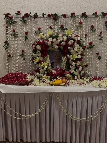 Another memorial setup with a beautiful floral wreath around the photograph. The table is decorated with a white cloth, scattered petals, and a garland of jasmine flowers.