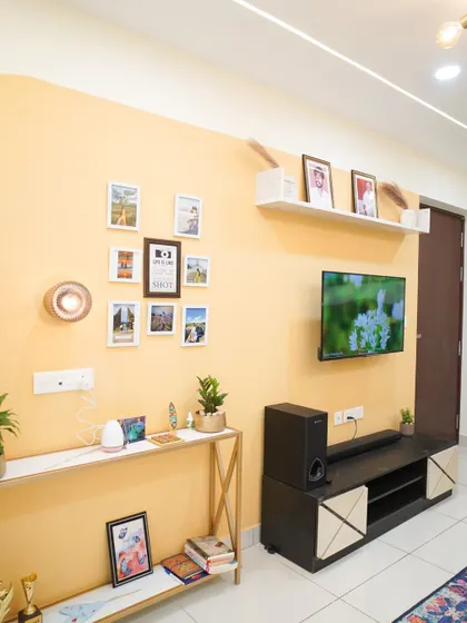 A living room with a bright yellow accent wall, a simple TV console, and a floating shelf for photos. The space is personalized with a gallery wall of family pictures.