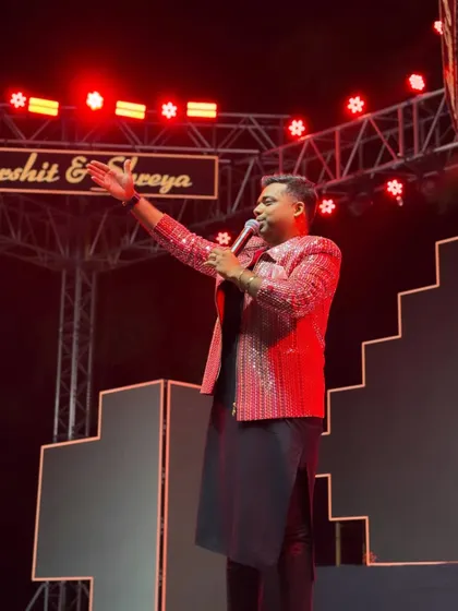 On stage, mic in hand, addressing the crowd during a lively Sangeet. The red stage lights reflect the high-energy atmosphere I love to create.