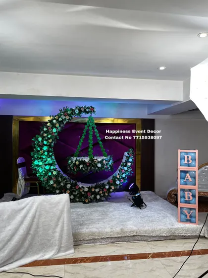 This elegant naming ceremony decor features a floral moon-gate with a hanging green cradle. The setup is completed with 'BABY' block letters and a clean white carpeted stage.