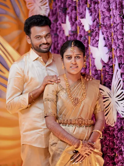A lovely portrait of a couple in matching gold outfits against a vibrant purple and gold butterfly-themed backdrop in Palladam.