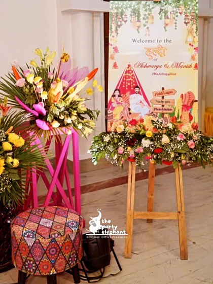 The welcome area for the Haldi ceremony, featuring a custom sign with traditional motifs, surrounded by vibrant floral arrangements and colorful printed stools.