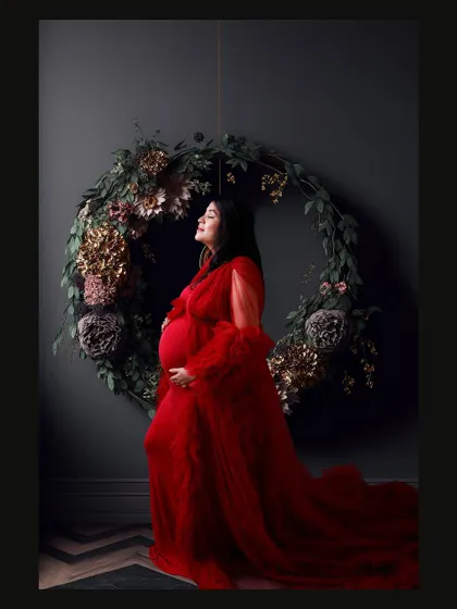 A side profile of a mom-to-be in a dramatic red gown, standing before a floral wreath. The dark, moody lighting creates a very artistic portrait.