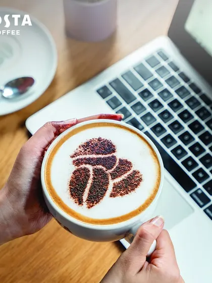 A person holds a cappuccino with coffee bean art over a laptop. This overhead shot perfectly combines the worlds of work and coffee, showing how I cater to the modern professional.