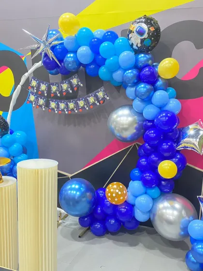 An outer space-themed birthday party with a crescent-shaped balloon arch in shades of blue and silver. The decor includes astronaut and star-shaped foil balloons.