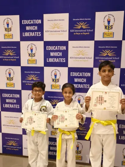 Three young karatekas from KES International School proudly display their new yellow belts and certificates. This is a significant first step in their martial arts training, marking their official entry into the world of karate.