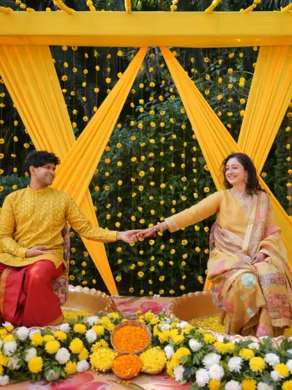 A happy couple shares a moment during their Haldi ceremony, seated in brass urlis filled with flower petals.