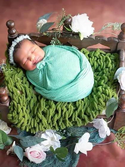 A sweet and serene portrait. The baby is wrapped in a soft mint green swaddle and rests on a tiny wooden bed, surrounded by white and green florals.