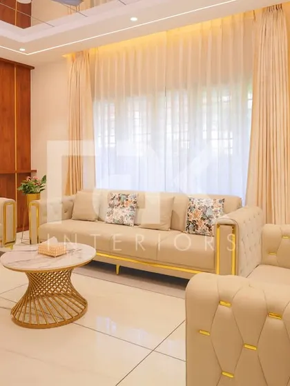 A luxurious living room with cream-colored tufted sofas featuring gold-plated legs. The mirrored wall panels and sheer curtains enhance the feeling of light and space.