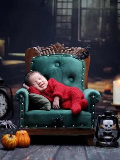 A spooky but sweet Halloween photoshoot. The baby is dressed in red and lounging on a miniature armchair in a haunted house setting with pumpkins and a skull lantern.